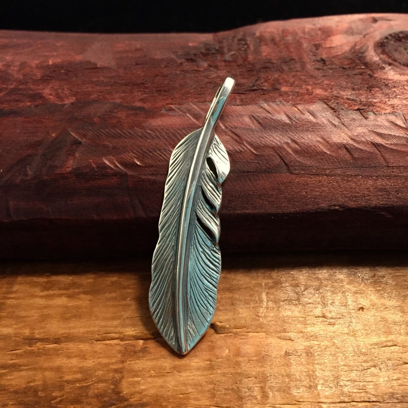 May include: A silver-colored feather pendant with detailed veining. The pendant is displayed against a dark wooden background, highlighting its intricate design. The feather's shape is elongated and slightly curved, suggesting a natural, organic form.