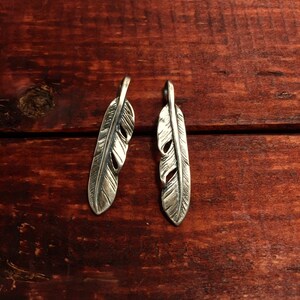 Puede incluir: Dos abalorios de plumas de plata con dise&ntilde;os detallados. Las plumas cuelgan de una superficie de madera.