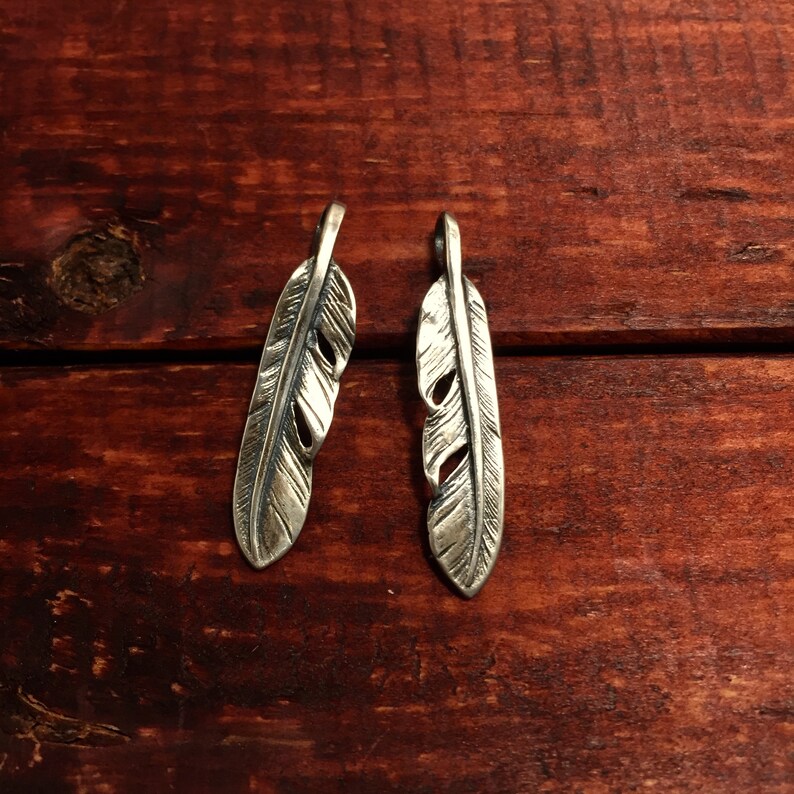 Puede incluir: Dos abalorios de plumas de plata con dise&ntilde;os detallados. Las plumas cuelgan de una superficie de madera.