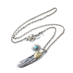 May include: A silver chain necklace with a silver and gold feather pendant. The pendant features a turquoise stone and a silver starburst design.