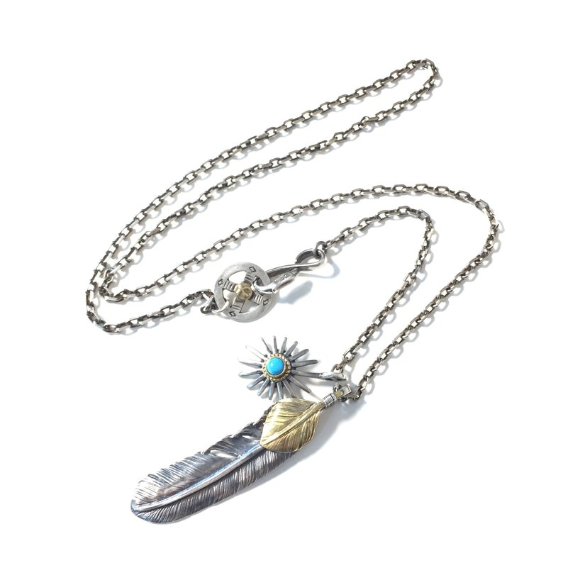 May include: A silver chain necklace with a silver and gold feather pendant. The pendant features a turquoise stone and a silver starburst design.