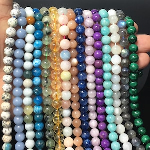 May include: A collection of 12 strands of gemstone beads, each strand featuring a different colour and type of stone. The beads are round and smooth, and they are arranged in a row. The colours of the beads include blue, green, purple, yellow, and white.