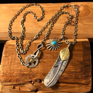 May include: A silver chain necklace with a silver and gold feather pendant, a silver starburst pendant with a turquoise stone, and a silver and brass clasp.