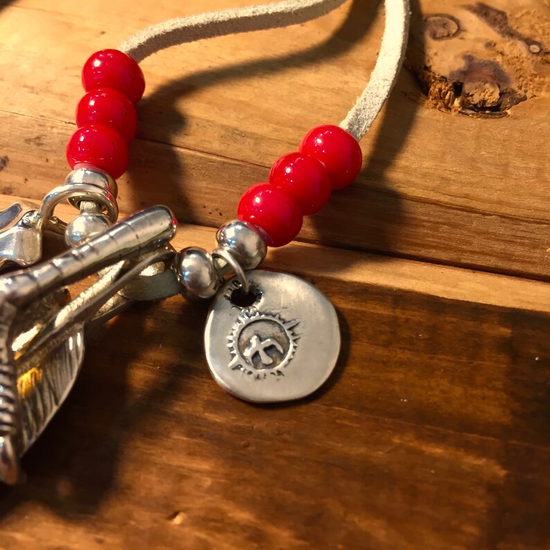 May include: A silver necklace with a charm featuring a stylized sun with a symbol inside. The necklace is made of suede cord and has red beads.
