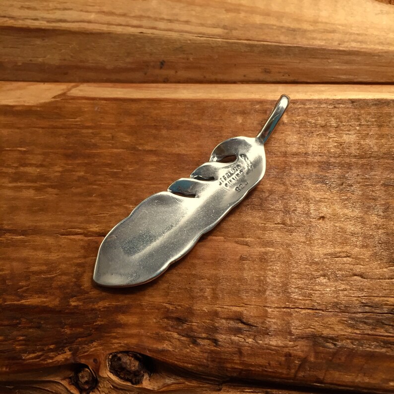 May include: A sterling silver feather pendant with a polished finish. The pendant has a curved shape with three cut-out sections and a loop at the top. The back of the pendant is stamped with "STERLING SILVER 925".