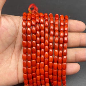 May include: A collection of bright orange-red barrel-shaped beads. The beads are strung together, with a red and yellow string visible at the top. The beads are smooth and evenly sized, creating a uniform pattern. The beads are held in a hand.