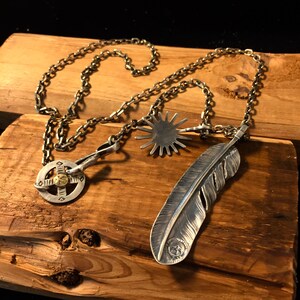 May include: A silver chain necklace with three pendants: a feather, a starburst, and a circular design with a gold center. The necklace is displayed on a wooden surface.