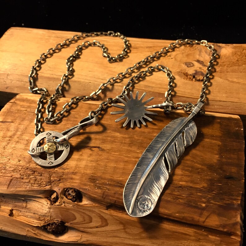 May include: A silver chain necklace with three pendants: a feather, a starburst, and a circular design with a gold center. The necklace is displayed on a wooden surface.