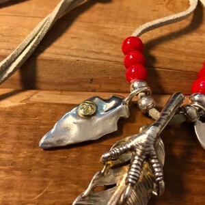May include: A silver necklace with a leather cord, red beads, and three silver charms: a feather with a claw, a silver arrowhead, and a round charm with a gold design.