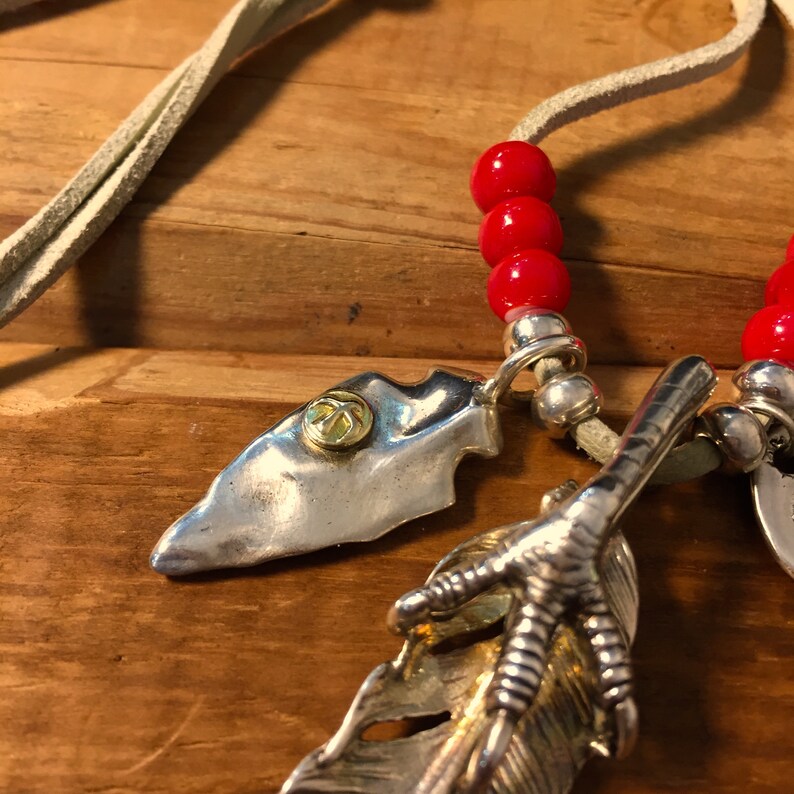 May include: A silver necklace with a leather cord, red beads, and three silver charms: a feather with a claw, a silver arrowhead, and a round charm with a gold design.