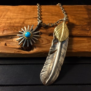 May include: A silver chain necklace with two pendants. One pendant is a silver feather with a gold overlay. The other pendant is a silver starburst with a turquoise stone in the center.