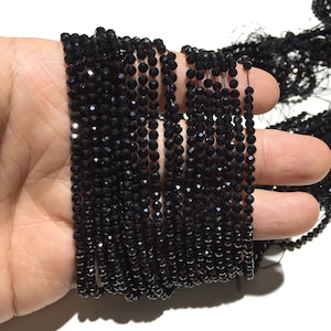 Puede incluir: Múltiples hebras de pequeñas cuentas negras facetadas. Las cuentas están ensartadas, creando múltiples líneas. Las cuentas se sostienen en una mano sobre un fondo blanco. Las cuentas tienen una superficie brillante y reflectante.