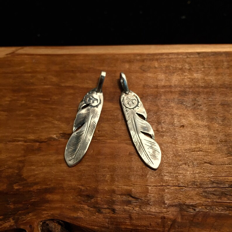 Puede incluir: Dos encantos de plumas de plata con un dise&ntilde;o detallado. Cada encanto tiene un c&iacute;rculo peque&ntilde;o con un s&iacute;mbolo en el centro.