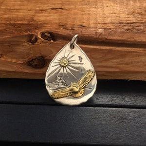 May include: A silver pendant shaped like a teardrop with a gold eagle in flight against a sunburst background.