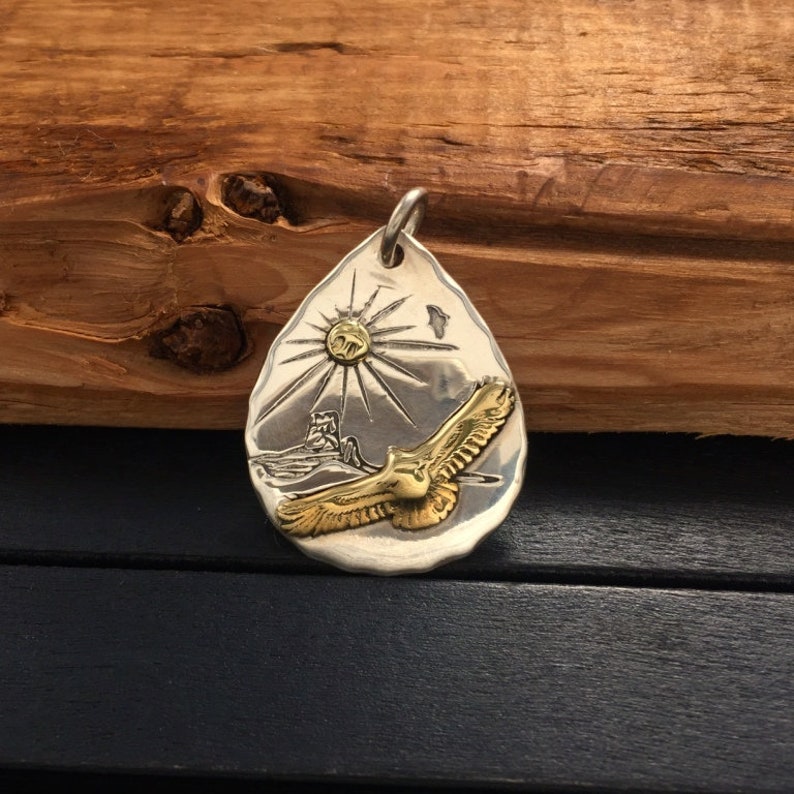 May include: A silver pendant shaped like a teardrop with a gold eagle in flight against a sunburst background.