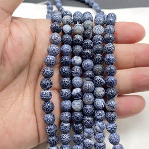 Pode incluir: Fios de contas redondas de ágata craquelada azul e branca. As contas têm uma cor de base azul escura com um padrão branco em forma de teia de aranha. As contas são enfiadas, prontas para fazer joias.