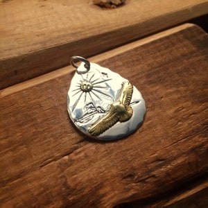 May include: A silver pendant with a textured surface depicting a sun and a golden eagle in flight. The pendant is shaped like a teardrop.