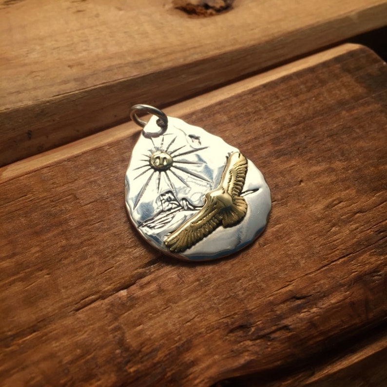 May include: A silver pendant with a textured surface depicting a sun and a golden eagle in flight. The pendant is shaped like a teardrop.
