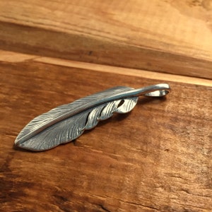 May include: A detailed silver feather pendant with intricate veining and a loop for a chain. The pendant is displayed on a wooden surface, showcasing its texture and design. This piece is ideal for jewelry.