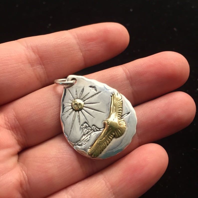 May include: Silver pendant with a gold eagle in flight, a sun, and mountains in the background. The pendant is shaped like a teardrop and has a textured surface.