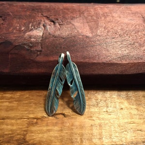 Puede incluir: Dos abalorios de plumas de plata con un acabado desgastado. Las plumas est&aacute;n detalladas con recortes y tienen una p&aacute;tina azul verdosa.