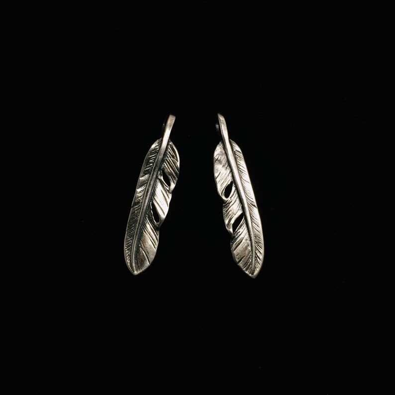 Puede incluir: Un par de pendientes de plata con forma de plumas. Las plumas son detalladas y tienen una superficie texturizada.