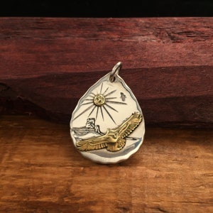 May include: A silver pendant with a gold eagle flying towards a sun with rays. The pendant is shaped like a teardrop and has a textured surface.