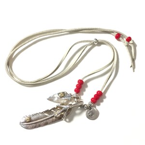 May include: A silver feather pendant necklace with a small arrowhead charm, a silver claw charm, and a round silver charm with a design. The necklace is on a light gray leather cord with red beads.