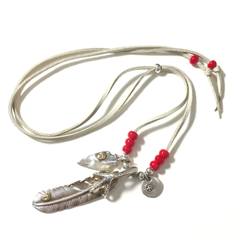 May include: A silver feather pendant necklace with a small arrowhead charm, a silver claw charm, and a round silver charm with a design. The necklace is on a light gray leather cord with red beads.