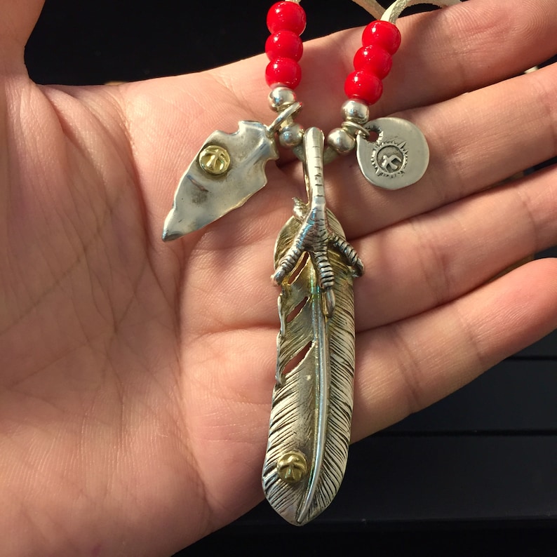 May include: Silver necklace with a feather pendant, a small arrowhead pendant, and a round pendant with a symbol. The necklace is made of red beads and a leather cord.