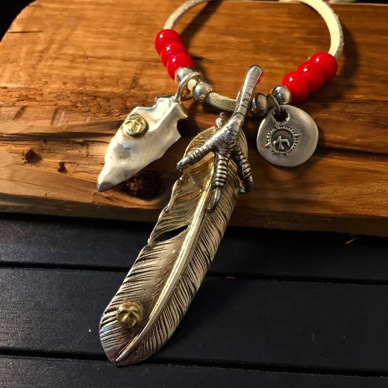 May include: A silver necklace with red beads, a silver arrowhead pendant, a silver feather pendant, and a silver charm with a symbol.