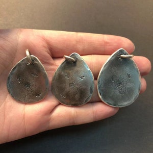 May include: Three silver teardrop-shaped pendants with a hammered texture and a small circle design in the center. Each pendant has a small loop at the top for attaching a chain.