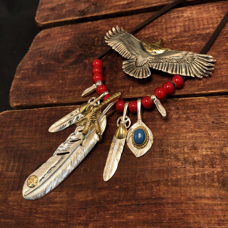 May include: A silver eagle pendant necklace with red beads and silver feather charms. The eagle is spread-winged and has a gold beak and talons. The feathers have a gold accent at the tip.