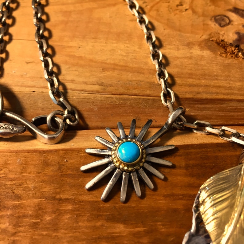 May include: A silver chain necklace with a turquoise stone pendant. The pendant is a starburst design with gold accents.