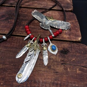 May include: A silver necklace with a red bead chain and multiple silver charms. The charms include a bird, a feather, and a claw. The necklace is on a wooden surface.