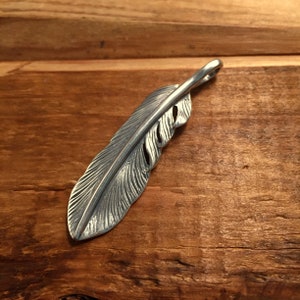 May include: A silver-colored feather pendant with intricate detailing. The pendant is designed to resemble a bird's feather, with a loop at the top for a chain. The feather is approximately 4 inches long.