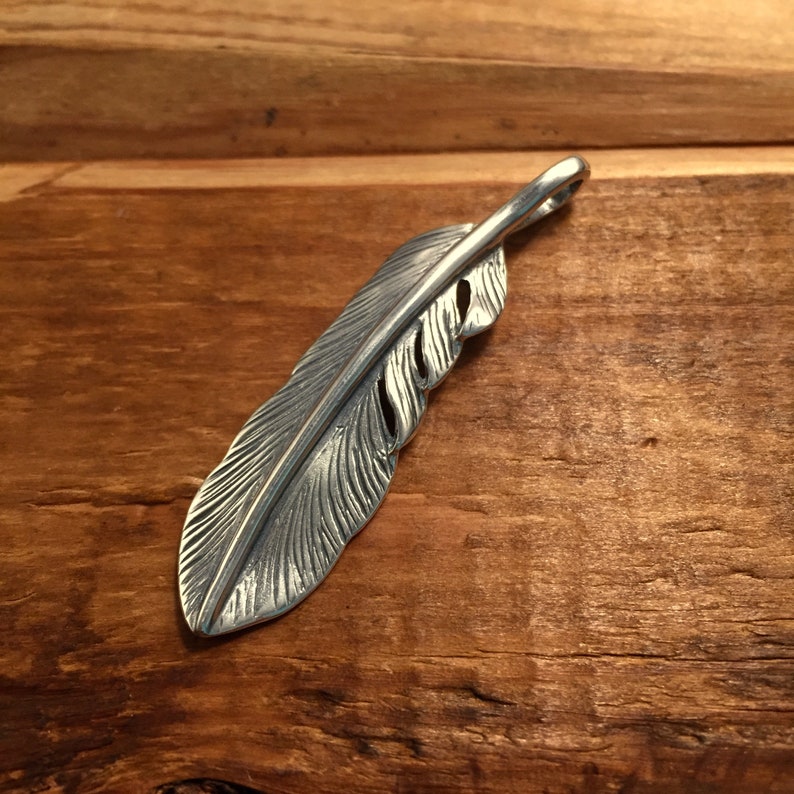 May include: A silver-colored feather pendant with intricate detailing. The pendant is designed to resemble a bird's feather, with a loop at the top for a chain. The feather is approximately 4 inches long.