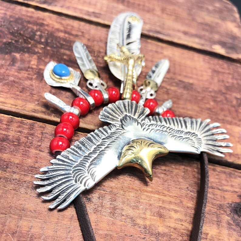 May include: A silver eagle pendant with gold accents, surrounded by red beads and silver feathers. The eagle has detailed wings and a beak.