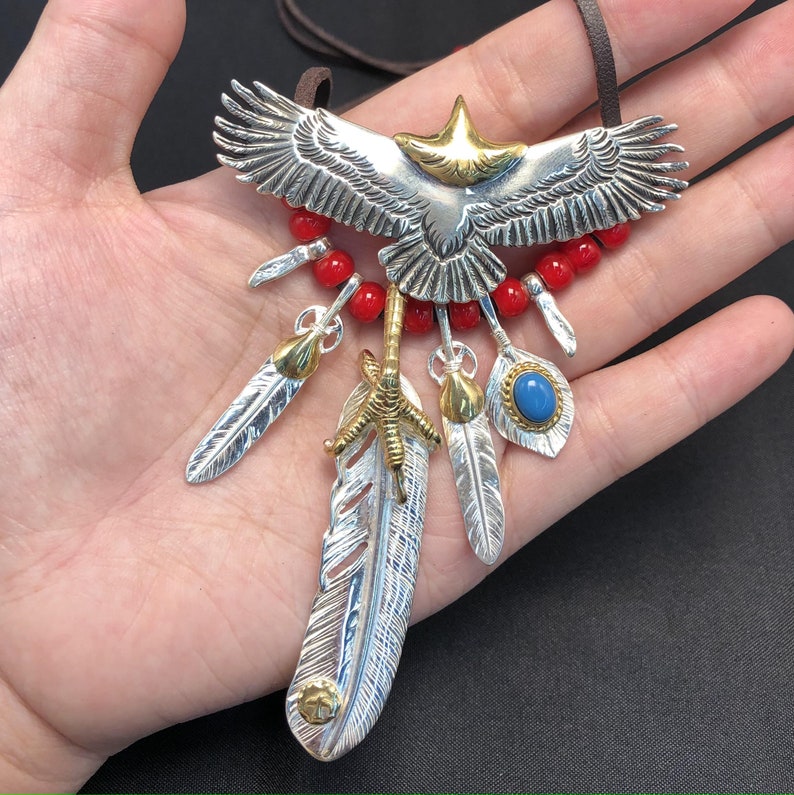 May include: Silver eagle pendant necklace with red beads, a blue stone, and multiple silver feathers. The eagle has a gold head and the pendant hangs from a brown leather cord.