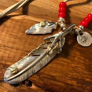 May include: A silver feather pendant necklace with a gold bird claw and a small arrowhead charm. The necklace is strung on a red beaded cord with a white leather cord.