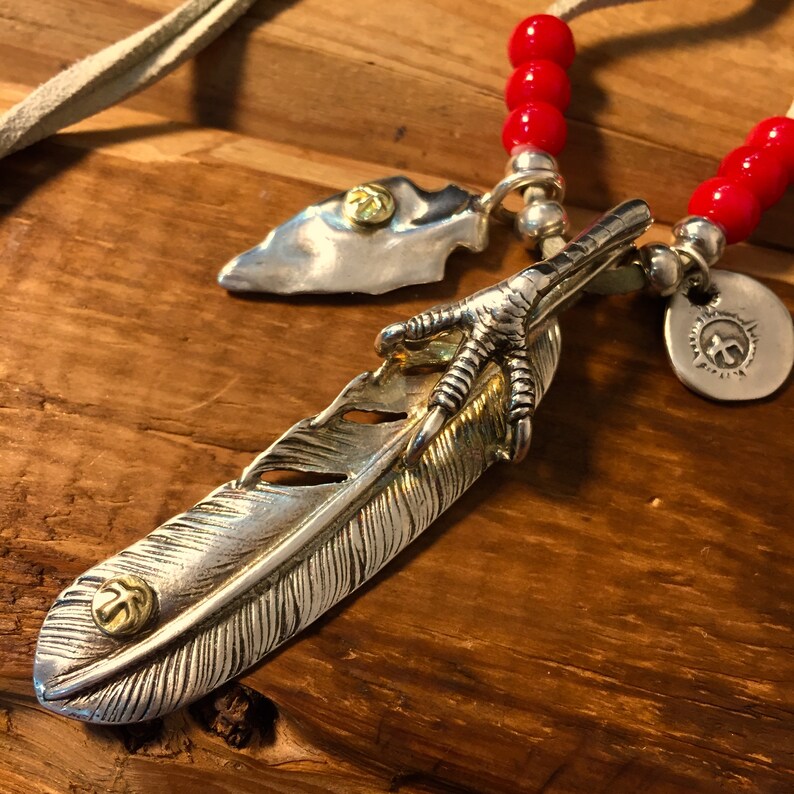 May include: A silver feather pendant necklace with a gold bird claw and a small arrowhead charm. The necklace is strung on a red beaded cord with a white leather cord.
