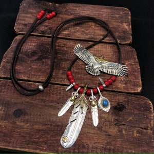 May include: A silver and gold necklace with a large eagle pendant and multiple feather charms. The necklace is made with red beads and a brown leather cord.