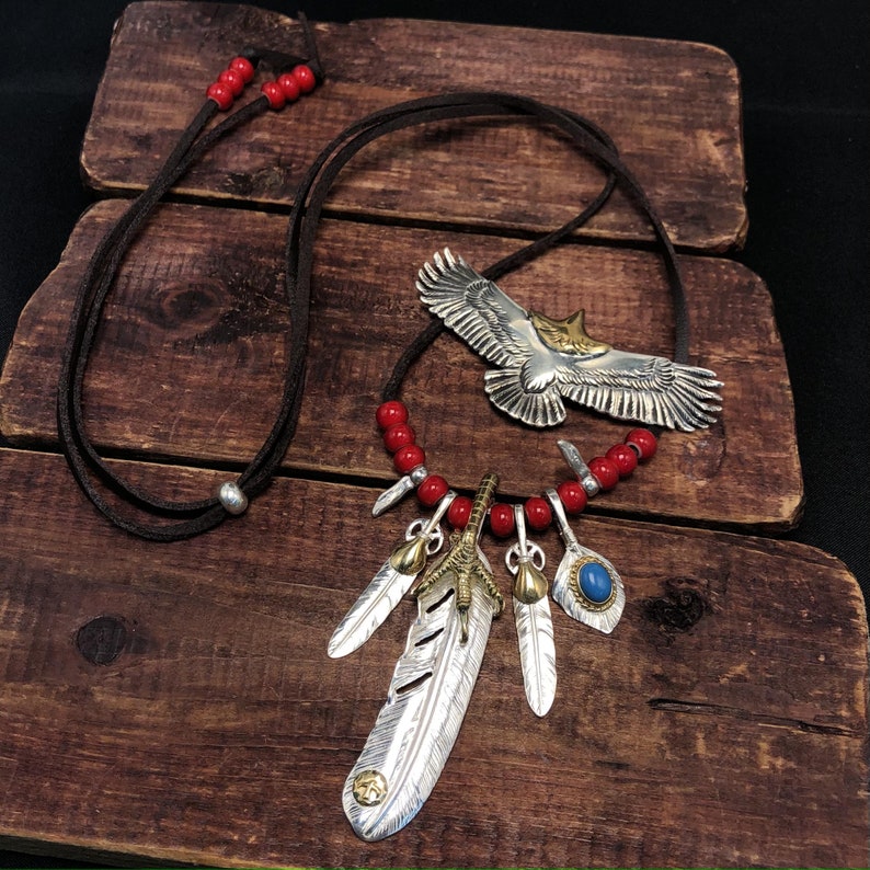 May include: A silver and gold necklace with a large eagle pendant and multiple feather charms. The necklace is made with red beads and a brown leather cord.
