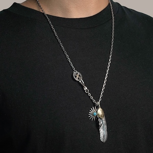 May include: A silver chain necklace with a silver and gold feather pendant. The feather has a turquoise stone at the top and a starburst design below it.