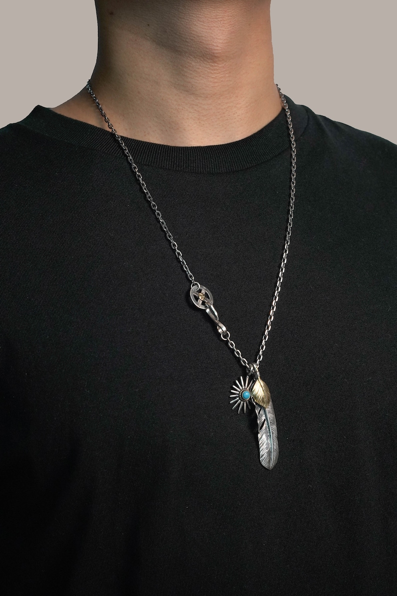 May include: A silver chain necklace with a silver and gold feather pendant. The feather has a turquoise stone at the top and a starburst design below it.