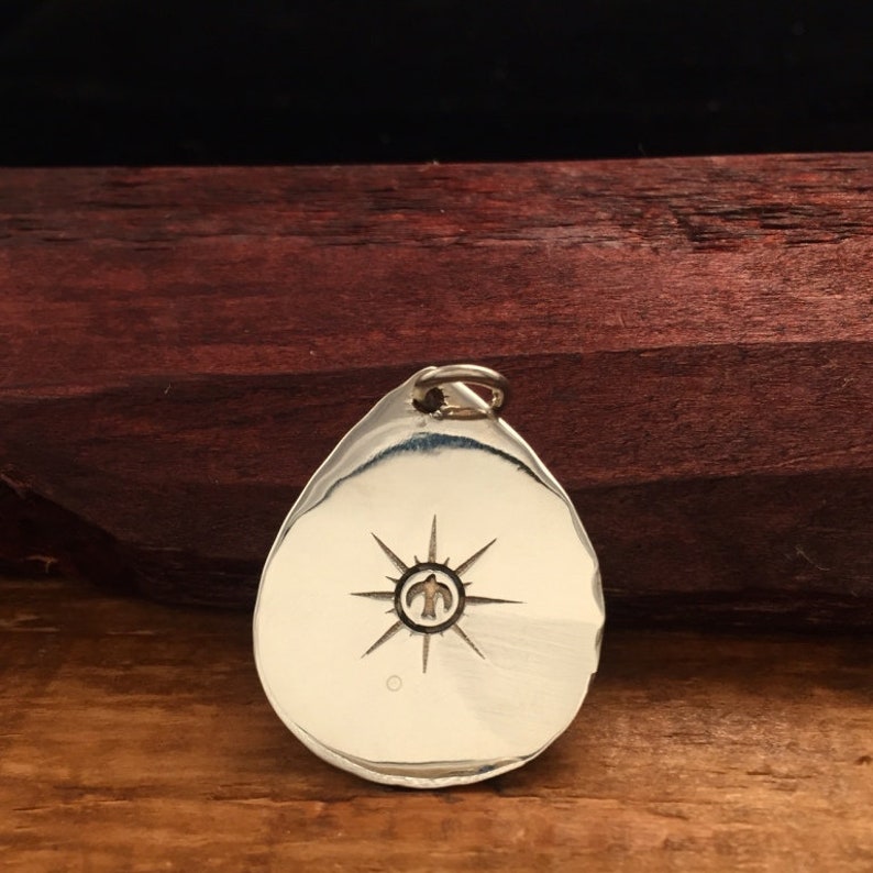 May include: A silver teardrop-shaped pendant with a starburst design and a small bird inside the star. The pendant has a loop at the top for attaching to a chain.
