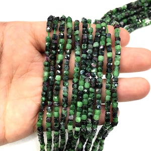 Może przedstawiać: Szereg zielonych i czarnych koralików z kamieni szlachetnych. Koraliki są fasetowane i mają kwadratowy kształt. Koraliki są nawleczone na cienką nić.