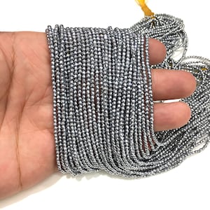 Könnte beinhalten: Eine Strähne silberfarbener Hämatitperlen mit einem Durchmesser von etwa 2 mm und einer facettierten Oberfläche. Die Perlen sind auf einem durchsichtigen Faden aufgefädelt und werden auf einer Hand gezeigt.