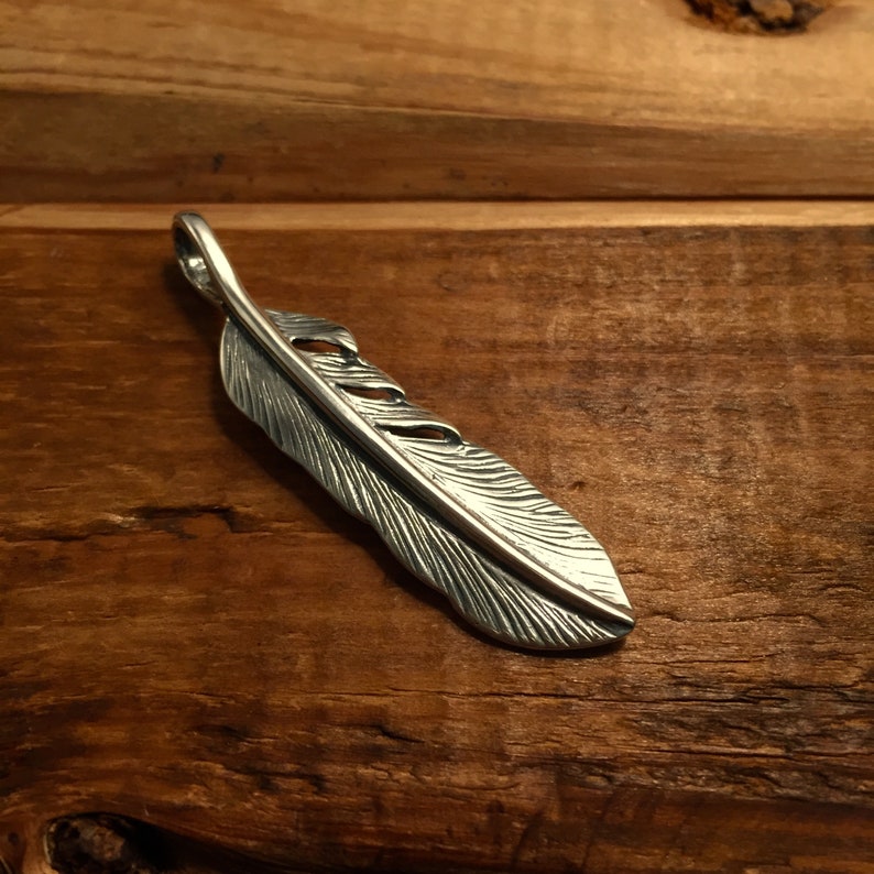 May include: A silver-colored feather pendant with intricate detailing. The pendant is designed to resemble a bird's feather, with a loop at the top for a chain. The feather's surface has detailed lines and textures, giving it a realistic appearance.