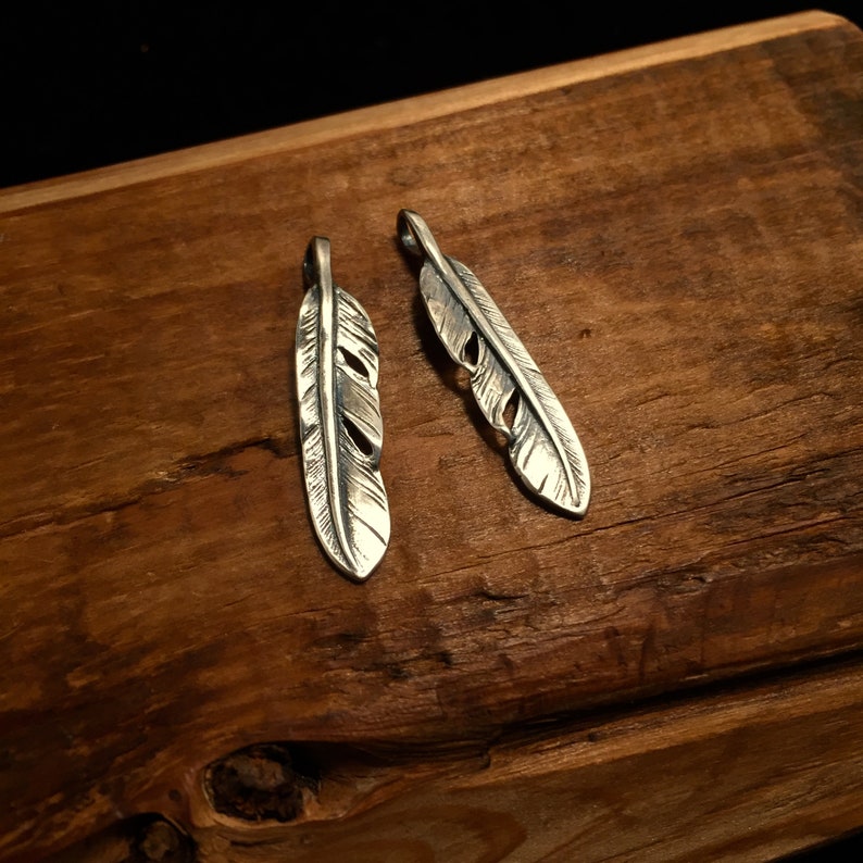 Puede incluir: Dos encantos de plumas de plata con grabado detallado. Las plumas cuelgan de una superficie de madera.